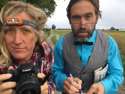 Greta i rutig skjorta har smuts på kinden och en pannlampa om huvudet och håller i en kamera. Dag har glasögon nere på näsan, grå väst på blå skjorta med svart fluga, och håller i penna och anteckningsblock.